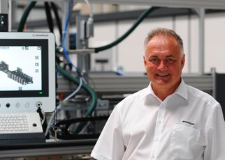Ein Mann in einem weißen Hemd steht lächelnd vor einem Bildschirm, der technische Informationen anzeigt, in einer industriellen Umgebung.