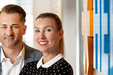 Drei Personen stehen in einem modernen Büro. Der Mann in einem Anzug und zwei Frauen, eine in einem schwarzen Rollkragenpullover und die andere in einem gepunkteten Oberteil.