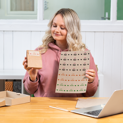 Eine Frau mit blonden, lockigen Haaren hält eine kleine Verpackung und ein Blatt mit Aufklebern in der Hand, während sie an einem Tisch sitzt.