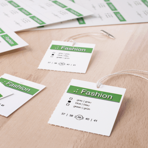 Modeetiketten mit Größenangaben und Farbauswahl in grünem und weißem Design auf einem Holzuntergrund.