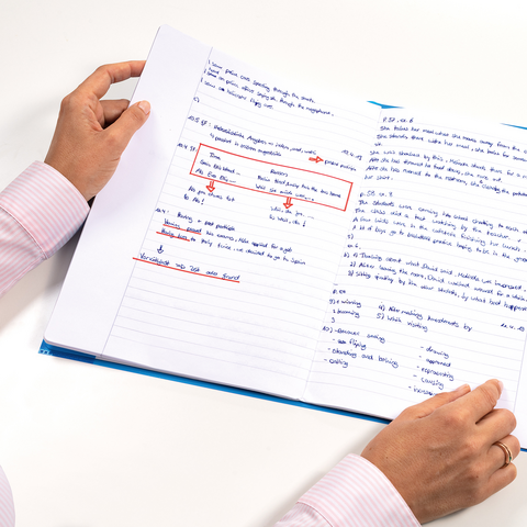 Eine Person hält ein aufgeschlagenes Notizbuch mit handschriftlichen Notizen und Markierungen in blauer und roter Tinte.