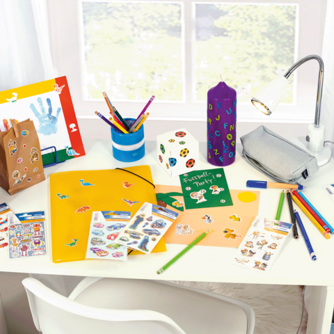 Bunt dekorierter Schreibtisch mit Bastelmaterialien, Aufklebern, Stiften, einem Ordner und einer Lampe.