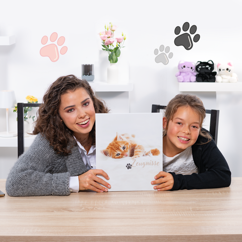 Eine Frau und ein Mädchen halten ein Buch mit einem Kätzchen-Cover, während sie an einem Tisch sitzen. Im Hintergrund sind dekorative Elemente sichtbar.