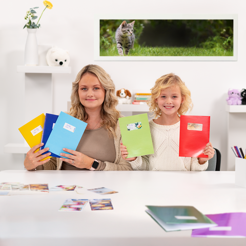 Eine Frau und ein Mädchen halten farbige Ordner in der Hand, während sie an einem Tisch mit Fotos sitzen. Im Hintergrund ist eine Katze zu sehen.