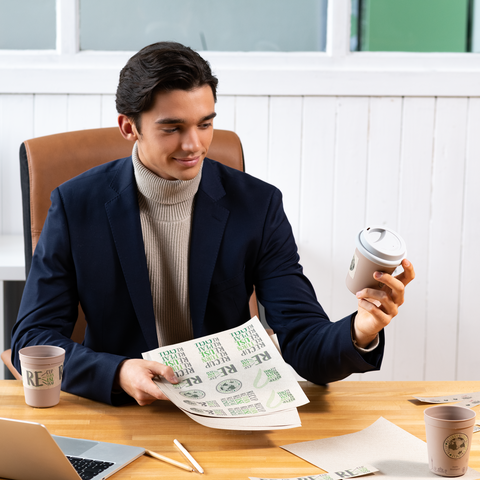 Ein junger Mann in einem Anzug hält einen Kaffeebecher und ein Blatt Papier mit Text in einer Büroumgebung.