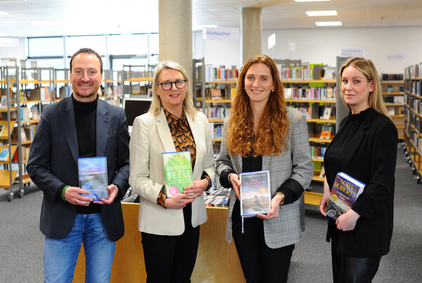 Vier Personen stehen in einer Bibliothek und halten Bücher in den Händen. Im Hintergrund sind Bücherregale sichtbar.