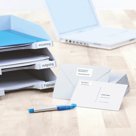 Ordner mit Beschriftungen für eingehende und ausgehende Dokumente, Umschläge und einen blauen Stift auf einem Tisch neben einem Laptop.