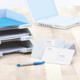Ordner mit Beschriftungen für eingehende und ausgehende Dokumente, Umschläge und einen blauen Stift auf einem Tisch neben einem Laptop.