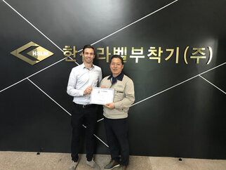 Zwei Männer halten ein Zertifikat vor einer Wand mit dem Schriftzug "HSLM" und koreanischen Zeichen.