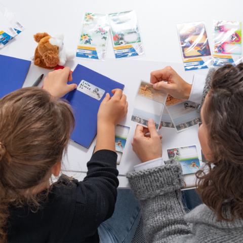 Eine Hand schreibt mit einem grünen Stift in ein Notizbuch, umgeben von bunten Aufklebern und einer Vase mit Blumen.