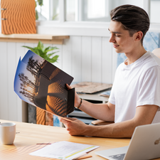 Ein junger Mann sitzt an einem Tisch und betrachtet eine Broschüre mit Bildern von Palmen und Sanddünen. Eine Tasse steht vor ihm.