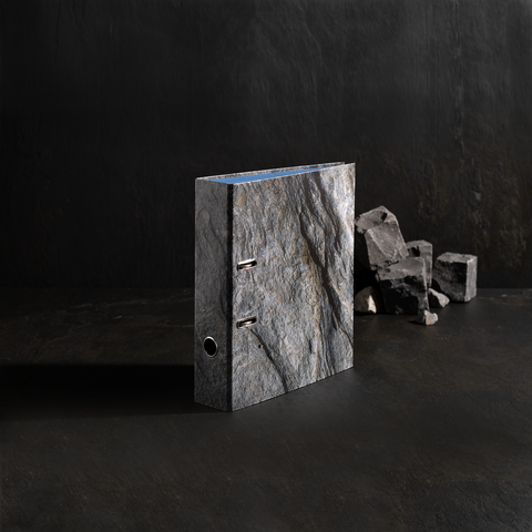 Ein grauer Ordner mit einer steinähnlichen Oberfläche steht auf einem dunklen Untergrund, neben einigen kleinen Steinen.