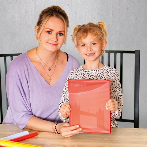 Eine Frau und ein kleines Mädchen sitzen an einem Tisch und halten ein rotes Heft mit der Aufschrift "English". Bunte Bastelmaterialien liegen auf dem Tisch.