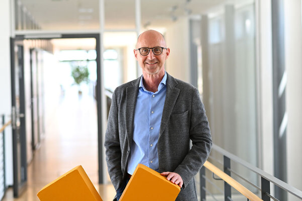 Ein Mann mit Brille steht in einem modernen Büroflur und hält zwei orangefarbene Kissen. Er trägt einen grauen Blazer und ein blaues Hemd.
