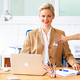 Eine Frau mit blonden Haaren sitzt an einem Schreibtisch, trägt einen Blazer und lächelt. Vor ihr steht ein Laptop und Schreibmaterial.