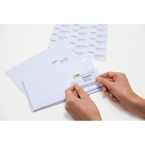 Eine Person hält einen adressierten Umschlag in der Hand, während mehrere leere Umschläge und ein Blatt mit Adressetiketten auf dem Tisch liegen.
