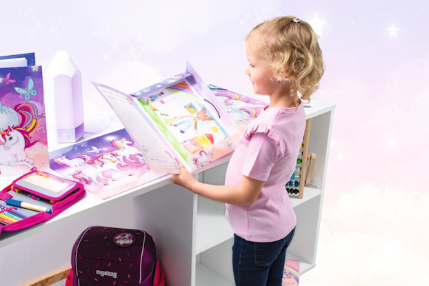 Ein Mädchen steht an einem Tisch und betrachtet die HERMA Einhorn Produkte von HERMA. Daneben liegen weitere Bücher und ein Rucksack.