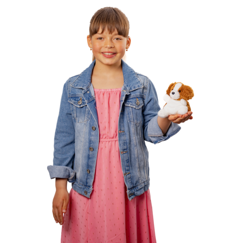 Ein Mädchen in einem rosa Kleid und einer Jeansjacke hält ein Stofftier in Form eines Hundes in der Hand und lächelt.