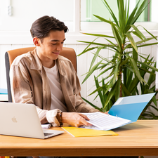 Ein junger Mann sitzt an einem Schreibtisch, arbeitet mit einem Laptop und blättert durch Dokumente in einer Ordner. Pflanzen und farbige Aktenordner sind im Hintergrund.