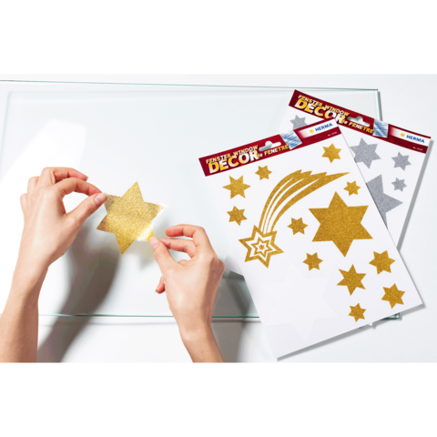 Eine Person platziert einen goldenen Sternaufkleber auf einer transparenten Oberfläche, umgeben von mehreren Bogen mit glitzernden Sternen.