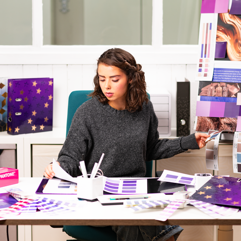 Eine Person sitzt an einem Tisch, umgeben von Farbmustern und Designmaterialien, während sie an einem kreativen Projekt arbeitet.