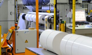 Industrielle Maschinen zur Verarbeitung von Rollenmaterial, mit einem Steuerpult im Hintergrund und einer Sicherheitsumzäunung.