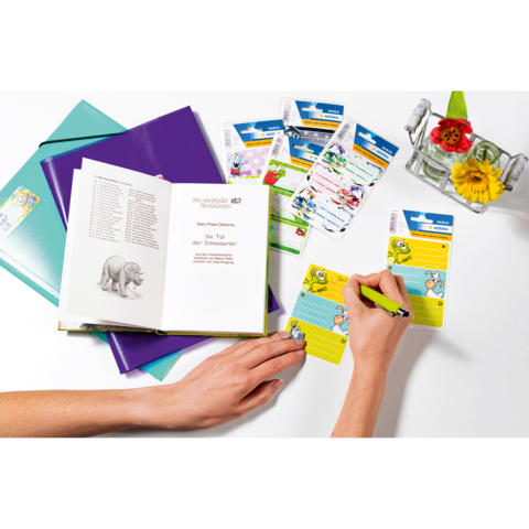 Eine Hand schreibt mit einem grünen Stift auf einer Karte, während ein offenes Buch und mehrere Karten im Hintergrund liegen.