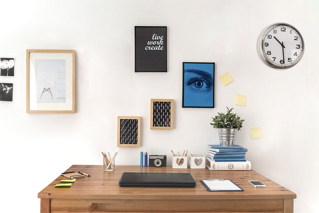 Ein Schreibtisch mit einem Laptop, dekorativen Bildern an der Wand, einer Pflanze und einer Stehlampe. Eine Wanduhr zeigt die Zeit an.