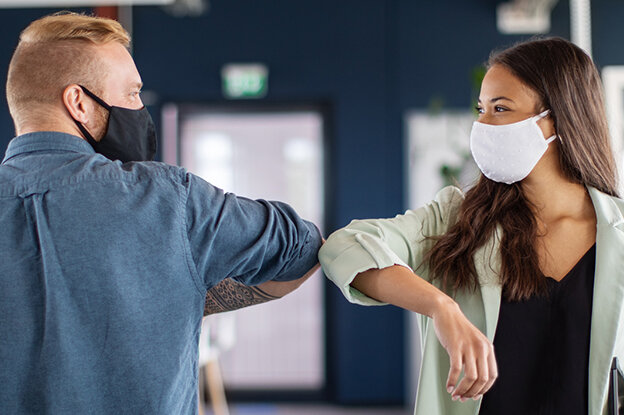 Zwei Personen in einem modernen Büro, die sich mit dem Ellenbogen begrüßen, beide tragen Gesichtsmasken.