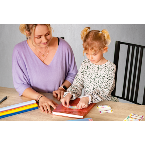Eine Frau und ein kleines Mädchen sitzen an einem Tisch, während sie gemeinsam an einem Buch arbeiten. Bunte Stifte und Karten liegen daneben.