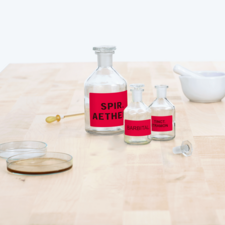 Flaschen mit roten Etiketten, beschriftet mit "Spir. Aether", "Barbital" und "Tinct. Tramon", auf einem Holztisch, daneben ein Mörser.