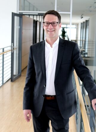 Ein Mann in einem Anzug steht in einem modernen Büroflur und lächelt, während er sich an einem Geländer festhält.