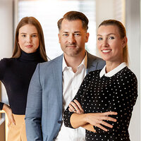 Drei Personen stehen in einem modernen Büro. Eine Frau trägt einen schwarzen Rollkragenpullover, ein Mann in einem Anzug und eine Frau in einem gepunkteten Oberteil.