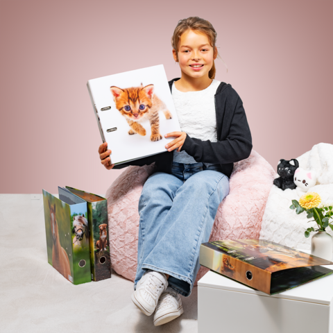 Ein Mädchen sitzt auf einem Sitzsack und hält einen Ordner mit einem Kätzchen auf dem Cover. Um sie herum sind weitere Ordner und eine Pflanze.
