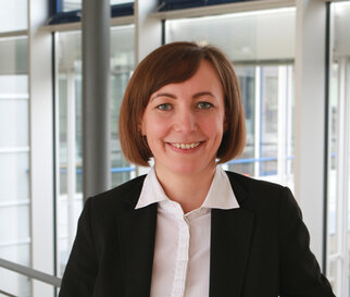 Eine lächelnde Frau mit schulterlangen, braunen Haaren trägt einen schwarzen Blazer und ein weißes Hemd. Sie steht in einem modernen Bürogebäude.