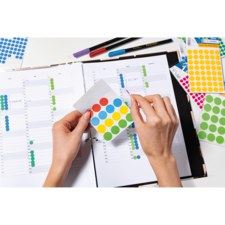 Eine Hand hält ein Blatt mit bunten Aufklebern über einem geöffneten Planer, der mit Wochenübersichten gefüllt ist.