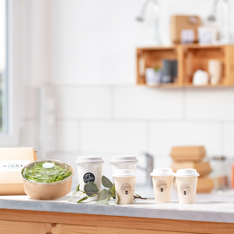 Ein Holztisch mit einer Schüssel Salat, mehreren Kaffeebechern und einer Verpackung, umgeben von einer hellen, modernen Küche.