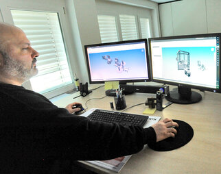 Ein Mann arbeitet an einem Computer mit zwei Bildschirmen, auf denen 3D-Modelle angezeigt werden. Der Schreibtisch ist mit Büromaterialien ausgestattet.