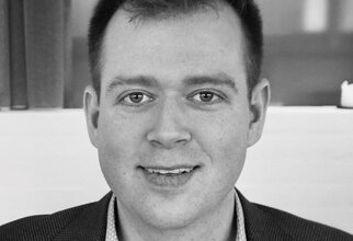 Ein Mann mit kurzem, dunklem Haar und einem freundlichen Lächeln, trägt einen Blazer und sitzt vor einem hellen Hintergrund.