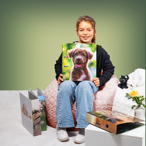 Ein Mädchen sitzt auf einem Pouf und hält einen Ordner mit einem Hundebild. Neben ihr liegen weitere Ordner und eine kleine Pflanze.