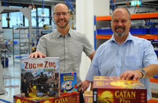 Zwei Männer stehen in einem Spielwarenlager und präsentieren Brettspiele, darunter "Zug um Zug Europa", "Halli Galli" und "Catan Plus".
