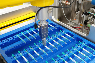 Ein automatisierter Roboterarm greift eine Spritze aus einem blauen Tray mit mehreren Spritzen, die ordentlich angeordnet sind.