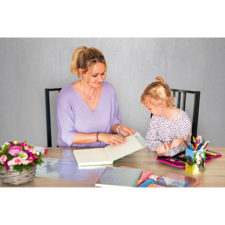 Eine Frau und ein Mädchen sitzen an einem Tisch und betrachten gemeinsam ein Buch. Auf dem Tisch liegen verschiedene Schreibmaterialien und Blumen.