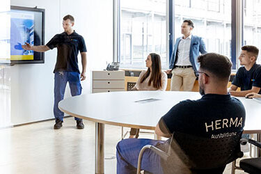 Ein Mann präsentiert an einem Bildschirm in einem modernen Besprechungsraum, während mehrere Personen am Tisch aufmerksam zuhören.