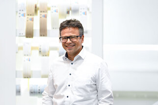 Ein Mann mit Brille und Hemd steht lächelnd vor einer hellen, modernen Hintergrundgestaltung mit verschiedenen Materialien.