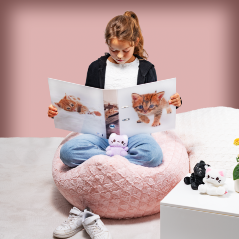 Ein Mädchen sitzt in einem großen, weichen Sitzsack und betrachtet ein Buch mit Bildern von Kätzchen. Plüschtiere liegen um sie herum.