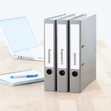 Drei graue Ordner mit Beschriftungen: Marketing, Controlling und Rechnungen, stehen auf einem Tisch neben einem Laptop und einem Notizblock.