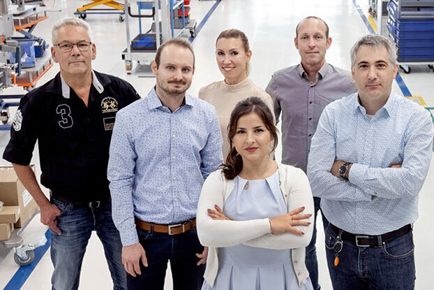 Eine Gruppe von sieben Personen steht in einer Fabrikhalle, umgeben von Maschinen und Arbeitsstationen.