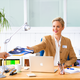 Eine Frau in einem Büro reicht einem Mann ein Dokument, während sie an einem Schreibtisch mit einem Laptop und Büromaterial sitzt.