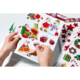 Hände halten ein Blatt mit bunten Weihnachtsstickern, darunter Weihnachtsmänner, Geschenke, einen Schneemann und Weihnachtsdekoration.